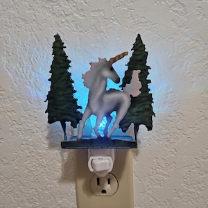 May include: A white unicorn night light with a blue glow, standing in a forest of two green trees. The night light plugs into a standard electrical outlet.