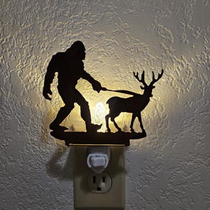 May include: A white night light with a silhouette of Bigfoot dragging a deer. The night light plugs into a standard electrical outlet.