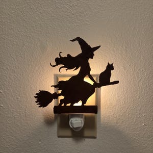 May include: A black silhouette night light featuring a witch wearing a pointed hat and riding a broomstick with a cat sitting behind her. The light emits a warm glow from behind the cutout design, which is mounted onto a standard wall outlet.