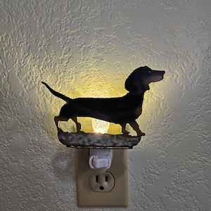 May include: A black dachshund silhouette night light plugged into an electrical outlet. The light is on and illuminating the dachshund's outline.