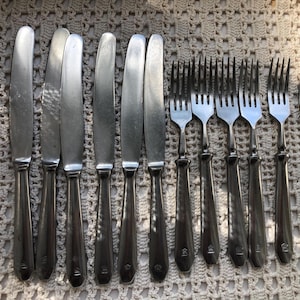 May include: A collection of vintage silverware, including knives and forks, arranged on a textured surface. The knives have rounded blades and the forks have four tines. The silverware is made of a shiny, silver-colored metal.