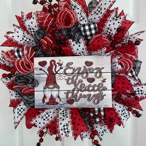May include: A decorative wreath with red, black, and white ribbons, berries, and a wooden sign. The sign reads "Enjoy the little things" and features a gnome illustration. The wreath is designed for home decor.
