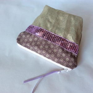 May include: A small, beige fabric pouch with a purple sequin trim. The pouch has a geometric pattern and a purple drawstring.