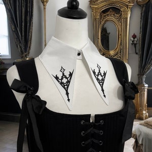Könnte beinhalten: Ein abnehmbarer weißer Kragen mit einem schwarzen gotischen Design. Der Kragen hat eine spitze Form und verfügt über einen Knopfverschluss.