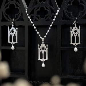 May include: A silver gothic-style jewelry set featuring a pendant necklace and matching earrings. Each piece has a cathedral window design and a dangling pearl. The jewelry is displayed against a black backdrop.