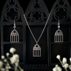 May include: A silver-toned jewelry set consisting of a pendant necklace and a pair of matching drop earrings. Each piece features a cutout design shaped like a pointed gothic arch window with intricate tracery and a central quatrefoil motif.