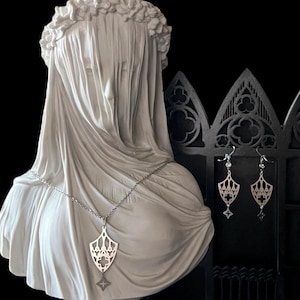 May include: A silver necklace and earrings set with a delicate, intricate design. The pendant and earrings feature a geometric, gothic-inspired shape with a cross at the bottom.