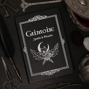Könnte beinhalten: Ein schwarzes, ledergebundenes Buch mit dem Titel "Grimoire Spells & Rituals" in weißen Lettern. Das Buch hat einen dekorativen Rahmen und eine weiße Illustration eines Halbmondes und Blumen.