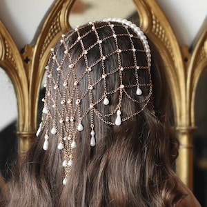 May include: A gold-toned headpiece with a net-like design, adorned with small pearls and dangling teardrop-shaped beads. The headpiece is worn over dark brown hair, with a pearl-lined band at the forehead. The background includes a gold-framed mirror.