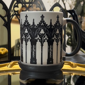 Puede incluir: Una taza de cerámica blanca con un diseño de arco gótico negro. La taza está sobre un posavasos negro y una bandeja dorada.