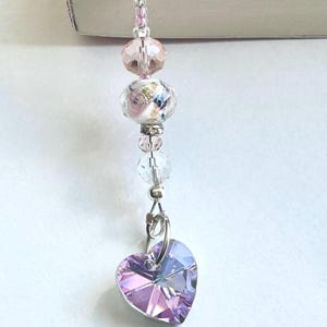 Puede incluir: Un marcapáginas decorativo con un dije de cristal en forma de corazón en tonos rosa y morado. El marcapáginas tiene cuentas rosas y transparentes, con una cuenta floral en el centro. Insertado en un libro.