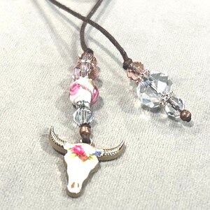May include: A handcrafted, Western-themed bookmark featuring a white bull skull pendant with floral accents. The pendant is suspended from a brown cord, adorned with clear and pink beads. The bookmark is designed for book lovers.