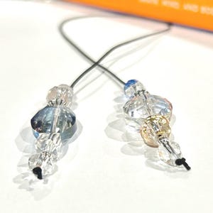 Op de afbeelding: Twee decoratieve kralenhangers met gefacetteerde heldere en blauw getinte kristallen kralen aan dunne zwarte koorden. Elke hanger bevat een grotere centrale kraal, kleinere accentkralen en metalen tussenstukjes, afgewerkt met een zwarte knoop.