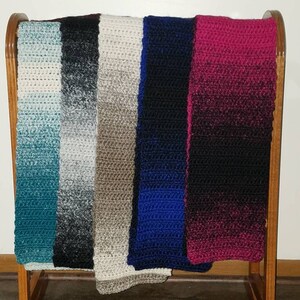 May include: Six crocheted scarves hanging on a wooden rack. The scarves are in various colors, including teal, black, beige, blue, and burgundy.
