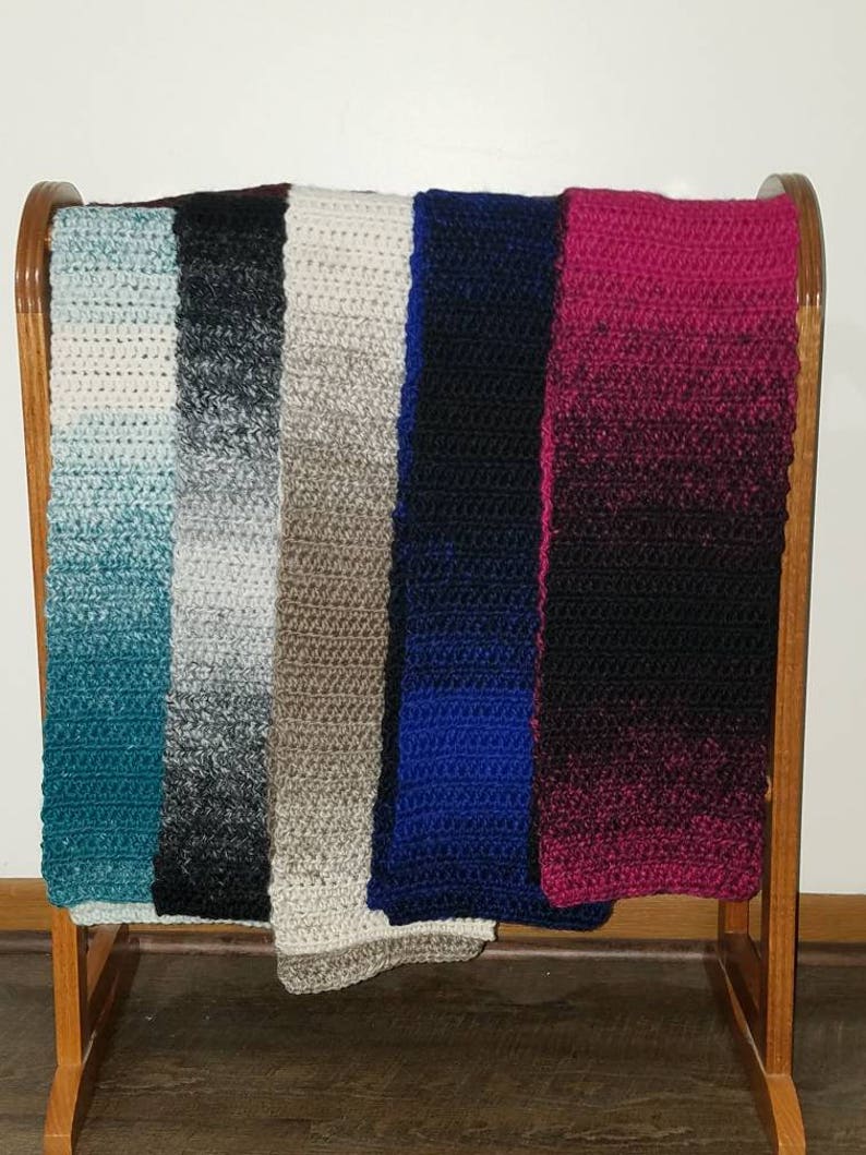 May include: Six crocheted scarves hanging on a wooden rack. The scarves are in various colors, including teal, black, beige, blue, and burgundy.