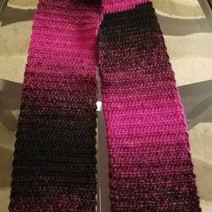 May include: A black and pink ombre crochet scarf. The scarf is made with a soft, textured yarn and has a gradient of color from black to pink.