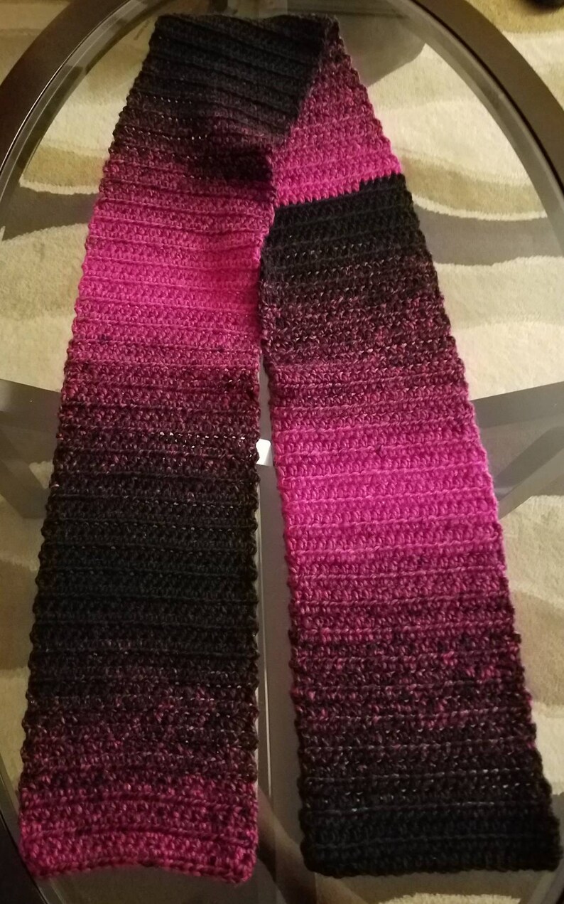 May include: A black and pink ombre crochet scarf. The scarf is made with a soft, textured yarn and has a gradient of color from black to pink.