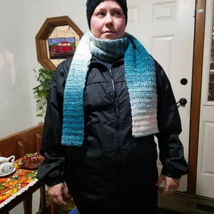 May include: A person wearing a black jacket, a black knit hat, and a blue and white ombre scarf. The scarf is crocheted and has a gradient of colors.
