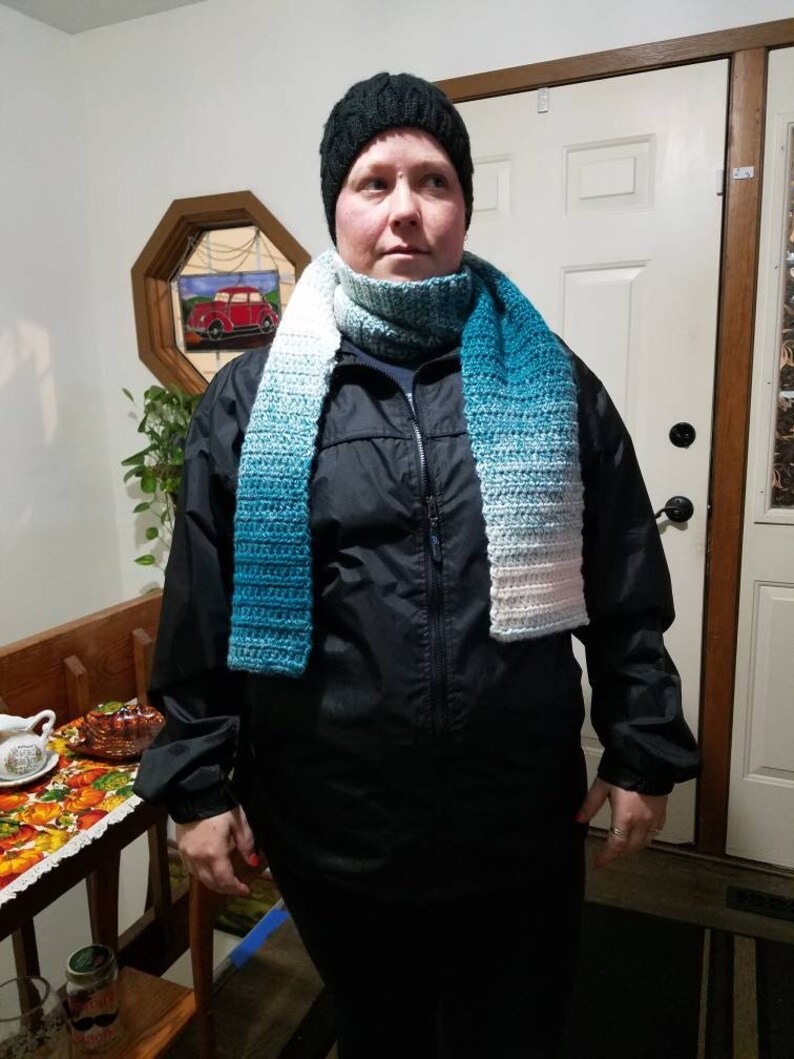 May include: A person wearing a black jacket, a black knit hat, and a blue and white ombre scarf. The scarf is crocheted and has a gradient of colors.