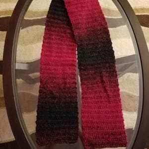 May include: A red and black ombre crochet scarf. The scarf is made with a soft yarn and has a subtle shimmer.