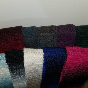 May include: A collection of ten crocheted scarves in various colors, including teal, black, white, blue, and pink. The scarves are displayed on a wooden rack.