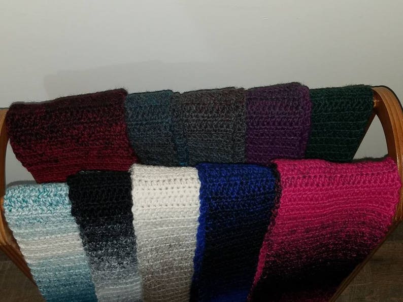 May include: A collection of ten crocheted scarves in various colors, including teal, black, white, blue, and pink. The scarves are displayed on a wooden rack.