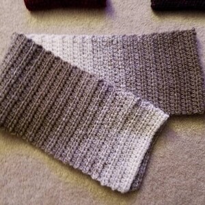 May include: A crocheted scarf with a gradient of beige and brown colors. The scarf is folded in half and the ends are visible.