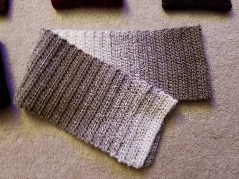 May include: A crocheted scarf with a gradient of beige and brown colors. The scarf is folded in half and the ends are visible.