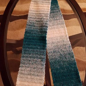 May include: A long, crocheted scarf with a gradient of teal, blue, and white colors. The scarf is folded in half and laid on a glass table.