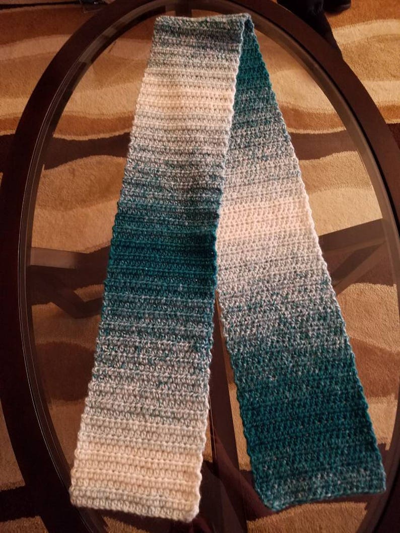May include: A long, crocheted scarf with a gradient of teal, blue, and white colors. The scarf is folded in half and laid on a glass table.