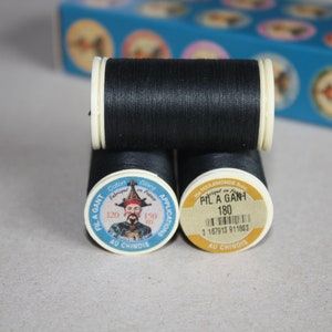 May include: Three spools of black thread with white and yellow labels. The labels feature text and a portrait of a man in a traditional hat. A blue box with circular designs is in the background.