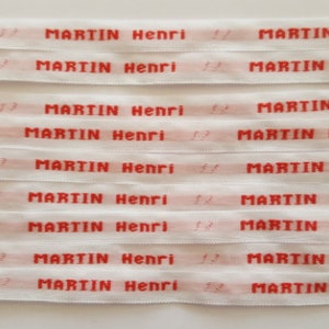 May include: Close-up of several white fabric ribbons with the name "MARTIN Henri" printed in red. The text is repeated across each ribbon. The ribbons are stacked horizontally.