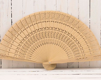 May include: A light brown wooden hand fan with intricate cut-out designs. The fan is closed and resting on a white wooden surface.
