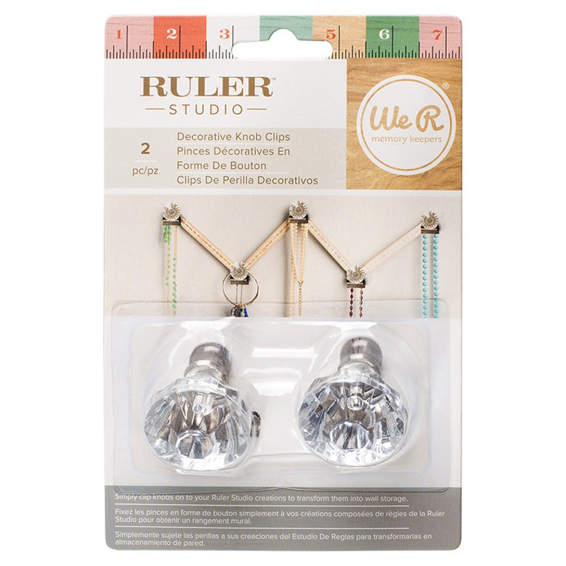 May include: Two clear plastic decorative knob clips with a diamond-shaped design. The clips are silver-toned and have a screw-on base. The packaging shows the clips being used to create a wall storage system. The packaging also includes the text "RULER STUDIO" and "We R memory keepers".