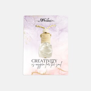 Puede incluir: Una botella de vidrio blanco con un tapón de madera que cuelga de una cuerda. La botella está sobre un fondo rosa y morado con purpurina dorada. El texto "CREATIVITY is oxygen for the soul" está impreso debajo de la botella.