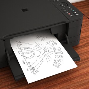 May include: A black and white printable coloring page featuring a mountain with a sun and the text "He is Risen". The page is being printed from a black printer.