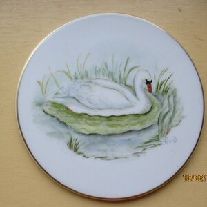 May include: A white porcelain plate with a gold rim features a hand-painted watercolor illustration of a white swan swimming in a pond with green reeds. The swan's head is turned to the side, and its beak is open slightly. The water is painted in shades of blue and gray, and the reeds are painted in shades of green. The plate is signed "G.W.D."