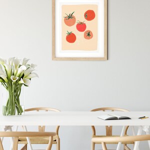 Op de afbeelding: Een ingelijst afdruk van een minimalistische illustratie van rode tomaten op een perzikkleurige achtergrond. De afdruk hangt aan een witte muur boven een witte tafel met vier houten stoelen.