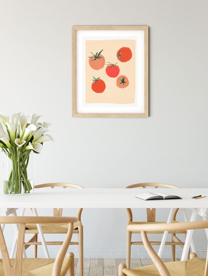 Op de afbeelding: Een ingelijst afdruk van een minimalistische illustratie van rode tomaten op een perzikkleurige achtergrond. De afdruk hangt aan een witte muur boven een witte tafel met vier houten stoelen.