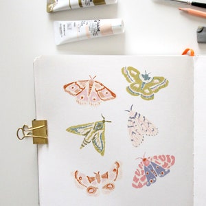 Puede incluir: Una p&aacute;gina de cuaderno de bocetos con seis ilustraciones de polillas pintadas a mano. Las polillas est&aacute;n pintadas en varios tonos de rosa, verde, azul y marr&oacute;n. El cuaderno de bocetos est&aacute; abierto y tiene un clip de carpeta dorado en el lado izquierdo.