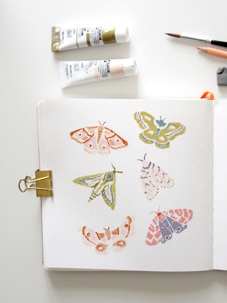 Puede incluir: Una p&aacute;gina de cuaderno de bocetos con seis ilustraciones de polillas pintadas a mano. Las polillas est&aacute;n pintadas en varios tonos de rosa, verde, azul y marr&oacute;n. El cuaderno de bocetos est&aacute; abierto y tiene un clip de carpeta dorado en el lado izquierdo.