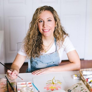 May include: A woman with curly blonde hair is painting a pink and yellow flower with watercolor paints. She is wearing a white t-shirt and a denim overall dress.