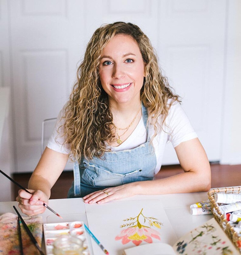 May include: A woman with curly blonde hair is painting a pink and yellow flower with watercolor paints. She is wearing a white t-shirt and a denim overall dress.