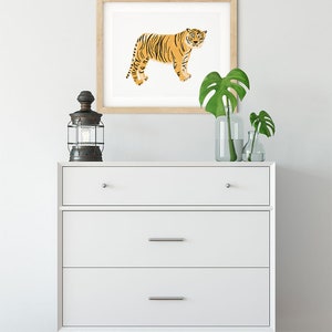 May include: A white chest of drawers with three drawers and wooden legs. A framed print of an orange tiger is hanging on the wall above the dresser. A black lantern and two potted plants are on the dresser.