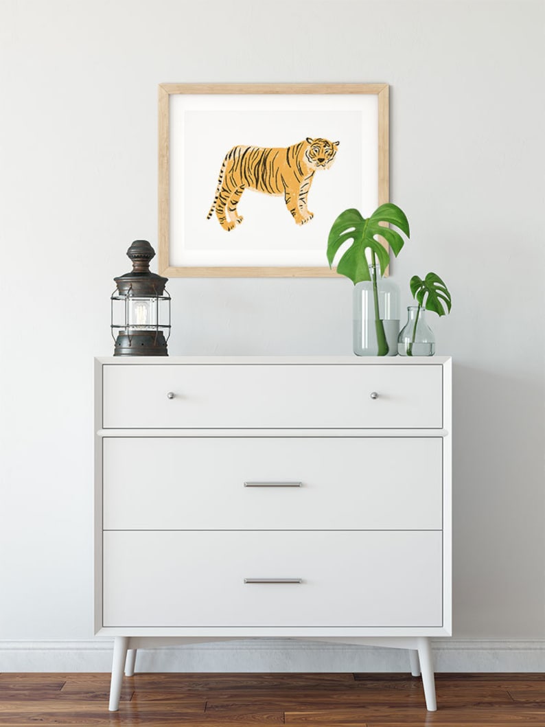 May include: A white chest of drawers with three drawers and wooden legs. A framed print of an orange tiger is hanging on the wall above the dresser. A black lantern and two potted plants are on the dresser.