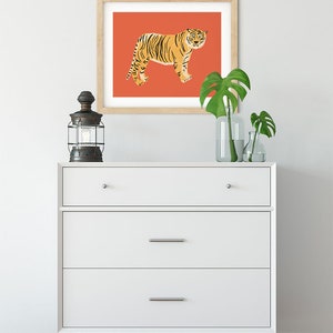May include: A white dresser with three drawers and wooden legs. A framed print of a tiger on a red background hangs above the dresser. Two potted plants are on the dresser.