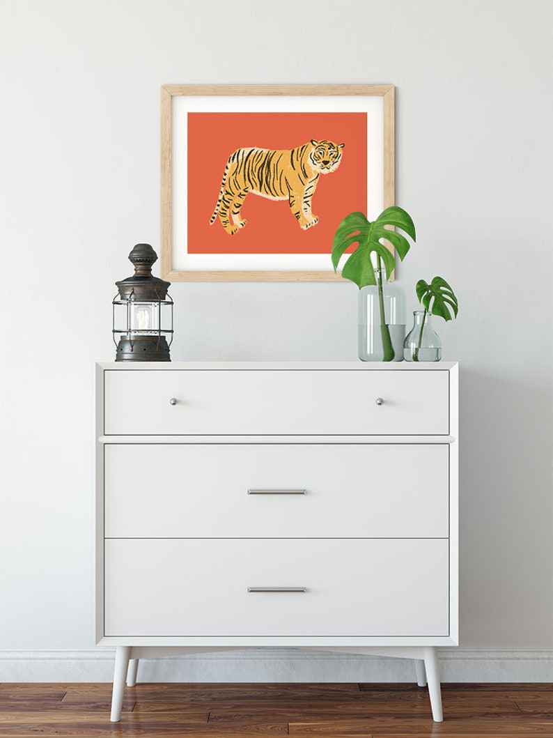 May include: A white dresser with three drawers and wooden legs. A framed print of a tiger on a red background hangs above the dresser. Two potted plants are on the dresser.