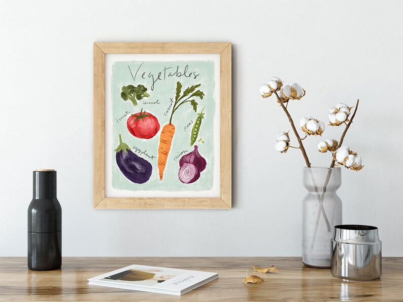 May include: A framed print with a light blue background featuring a watercolor illustration of various vegetables, including a tomato, carrot, onion, eggplant, broccoli, and peas. The text "Vegetables" is written in black cursive at the top of the print.
