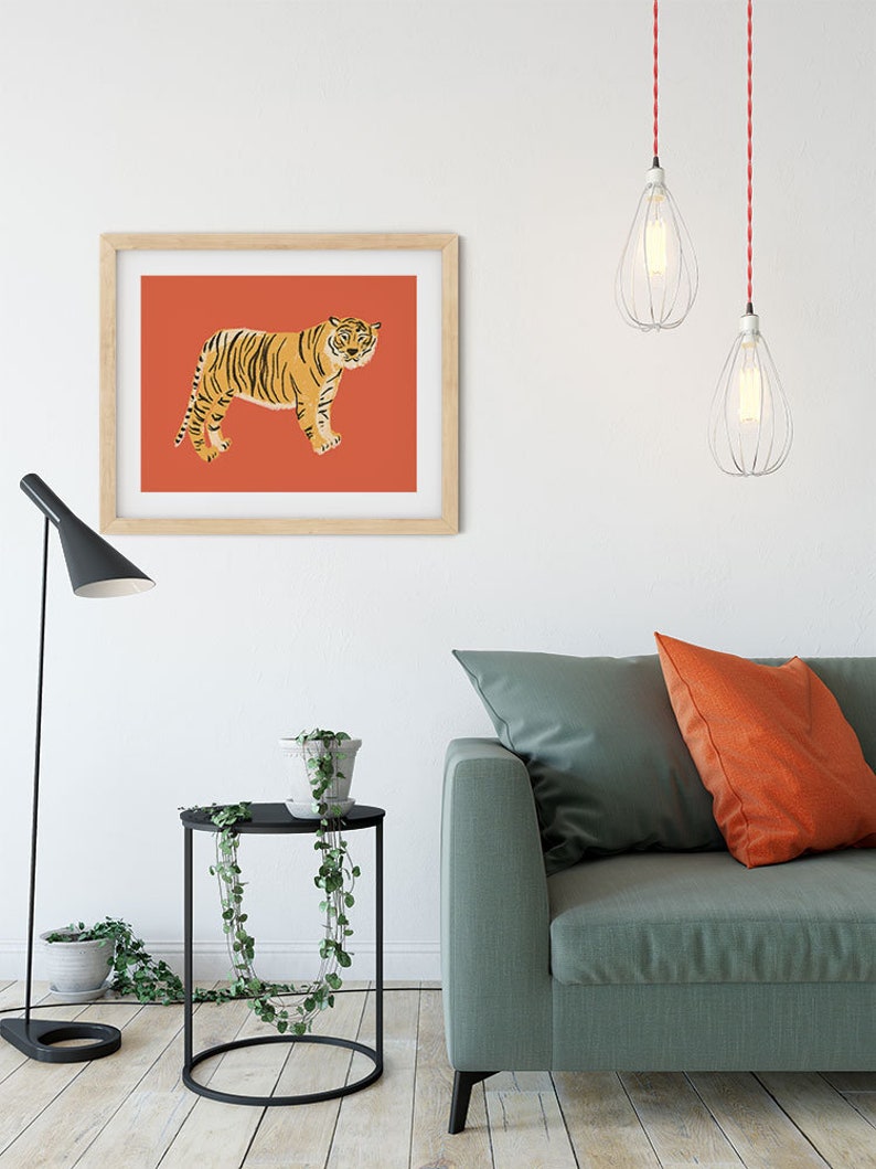 May include: A framed print of a tiger on a red background. The print is hanging on a white wall above a green sofa with orange throw pillows. There is a black side table with a potted plant next to the sofa.