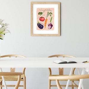 Puede incluir: Una impresi&oacute;n enmarcada con un fondo rosa y la palabra "Vegetales" escrita en cursiva negra. La impresi&oacute;n presenta una ilustraci&oacute;n de acuarela de un tomate, una zanahoria, br&oacute;coli, berenjena, cebolla y una vaina de guisante.